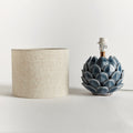 Decorative blue ceramic artichoke-shaped lamp on a white surface with a neutral-colored lampshade.