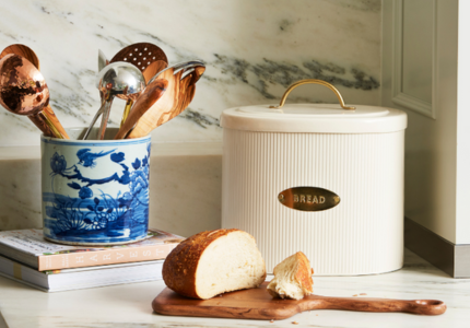 Bread bin with a loaf of bread on a wooden cutting board, surrounded by kitchen utensils.