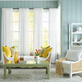 A styled living room interior featuring white sheer curtains hanging in front of a tall window with light blue paneled walls. The room includes a pale green sideboard with a mirror and lamp, a matching green coffee table, yellow accent pillows, a white armchair with a patterned throw, and a white shelving unit.