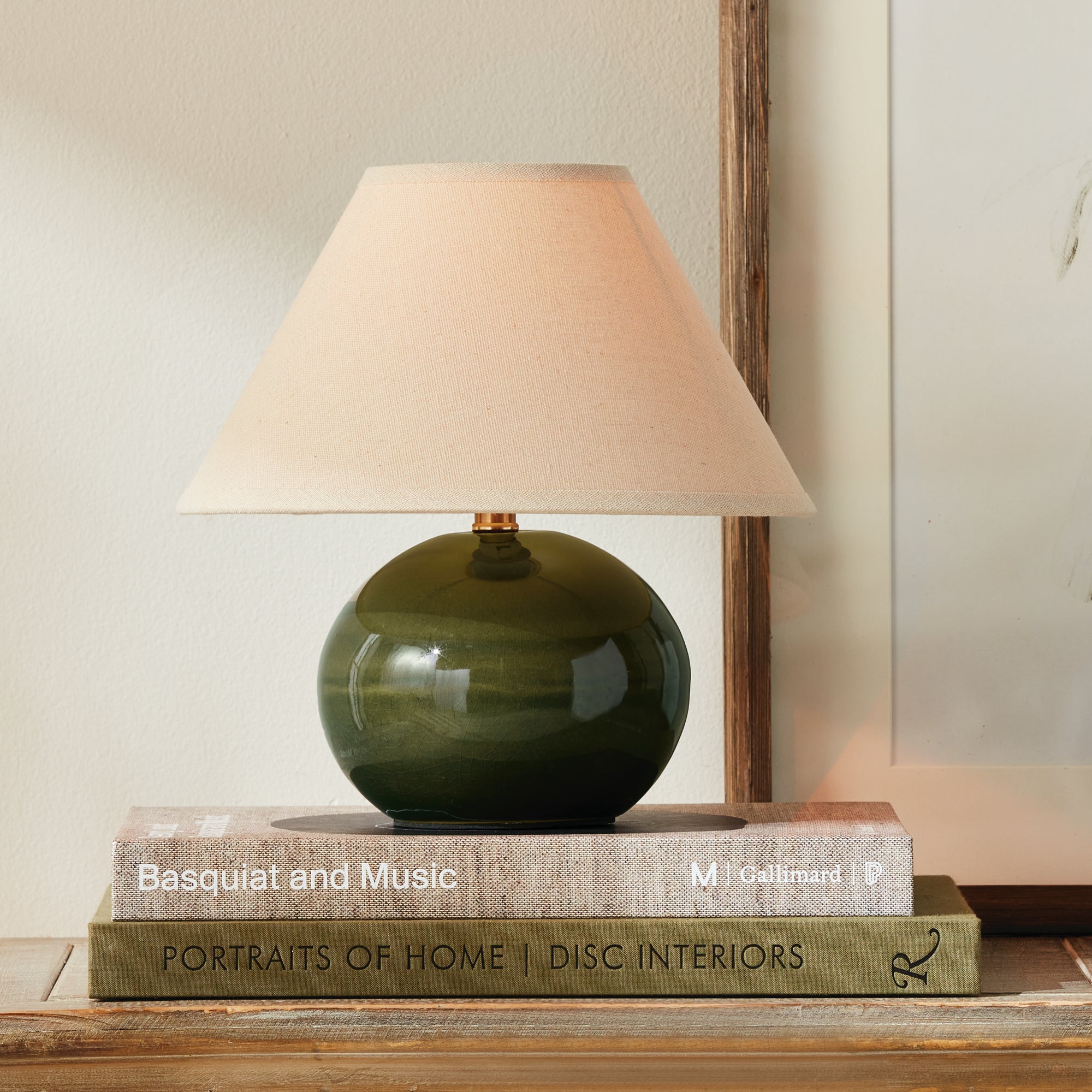 Green ceramic small lamp with beige lampshade on top of books on a wooden surface.
