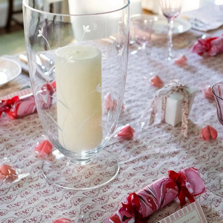 Decorative table setting with a candle in a glass holder, pink paper crackers, and a patterned tablecloth.