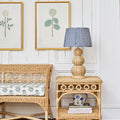 Rattan side table with a lamp and decor items against a white paneled wall with framed artwork.