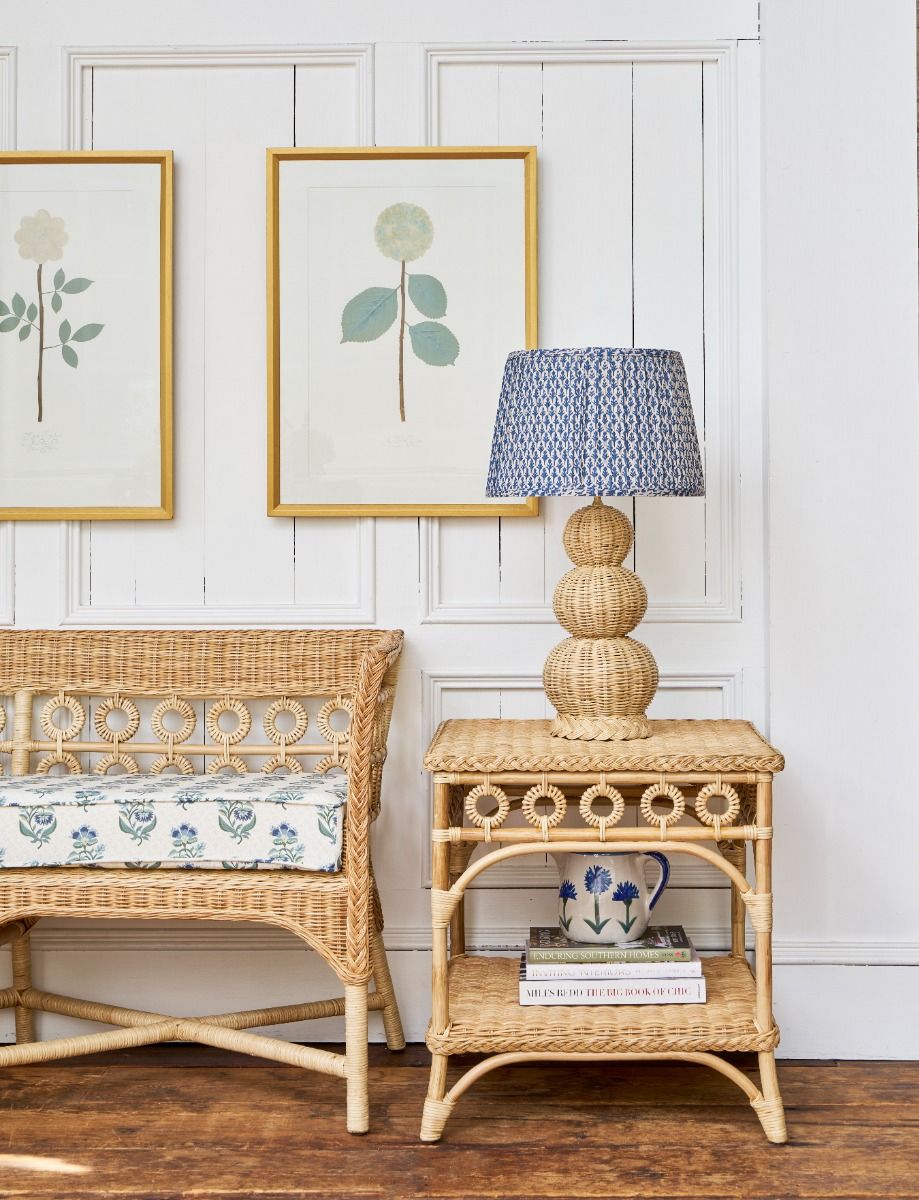 Rattan side table with a lamp and decor items against a white paneled wall with framed artwork.